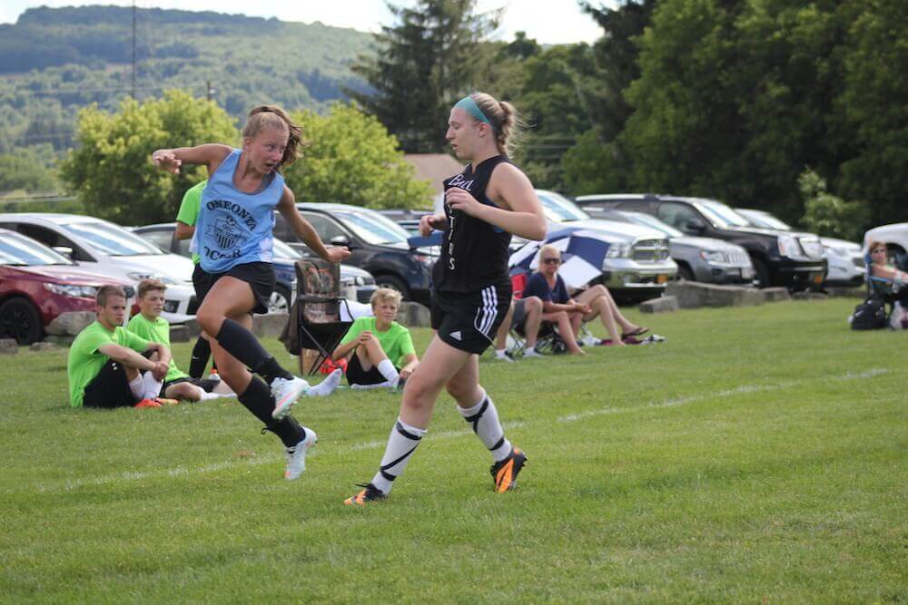 One girl in an OSC blue uniform jumps just after kicking the ball as an opposing player persues