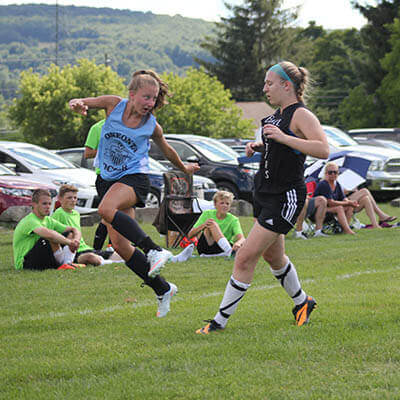 One girl in an OSC blue uniform jumps just after kicking the ball as an opposing player persues