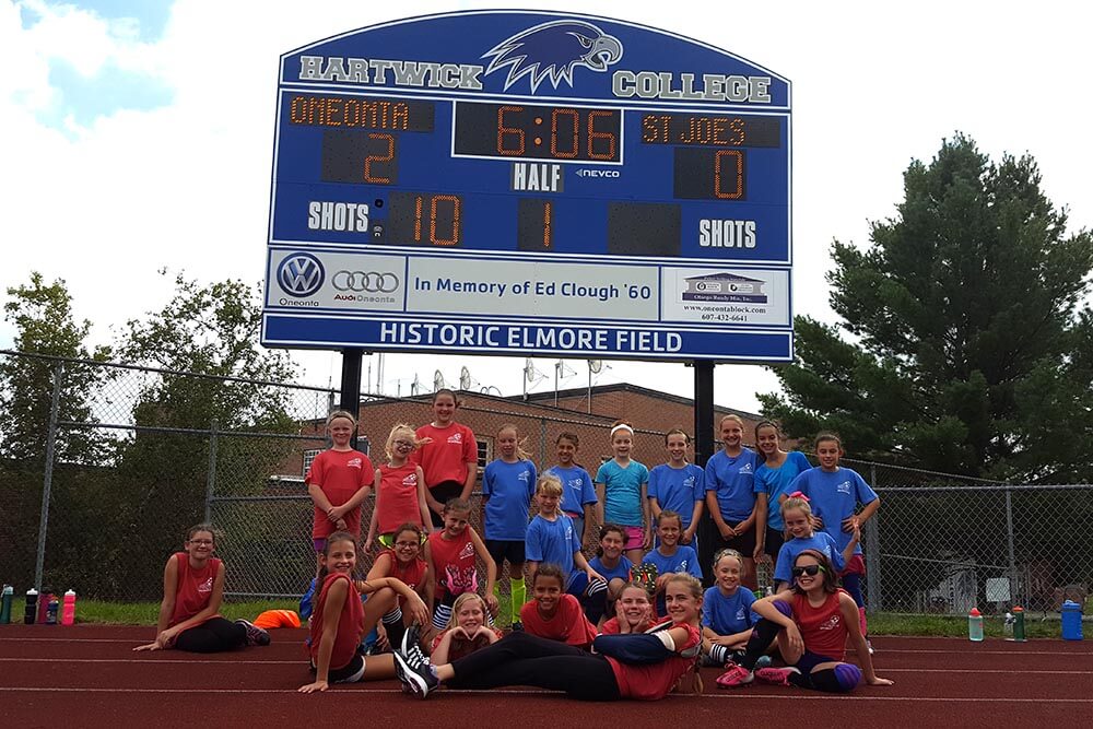 Players pose in front of the large, blue Hartwick scoreboard. The score reads two to zero Oneonta.